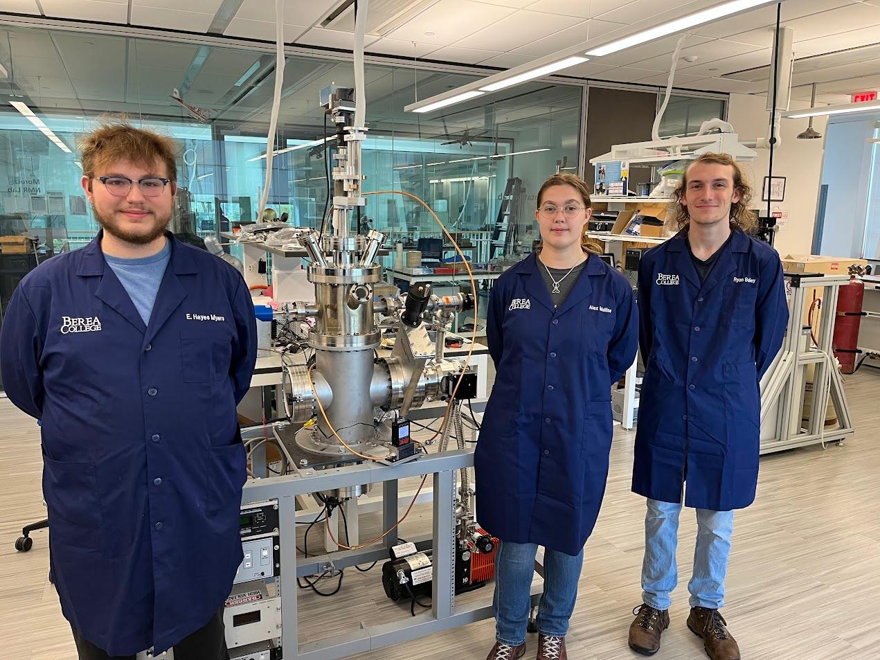 Three Berea College physics students wearing personalized lab coats stand beside a large machine in a lab classroom.
