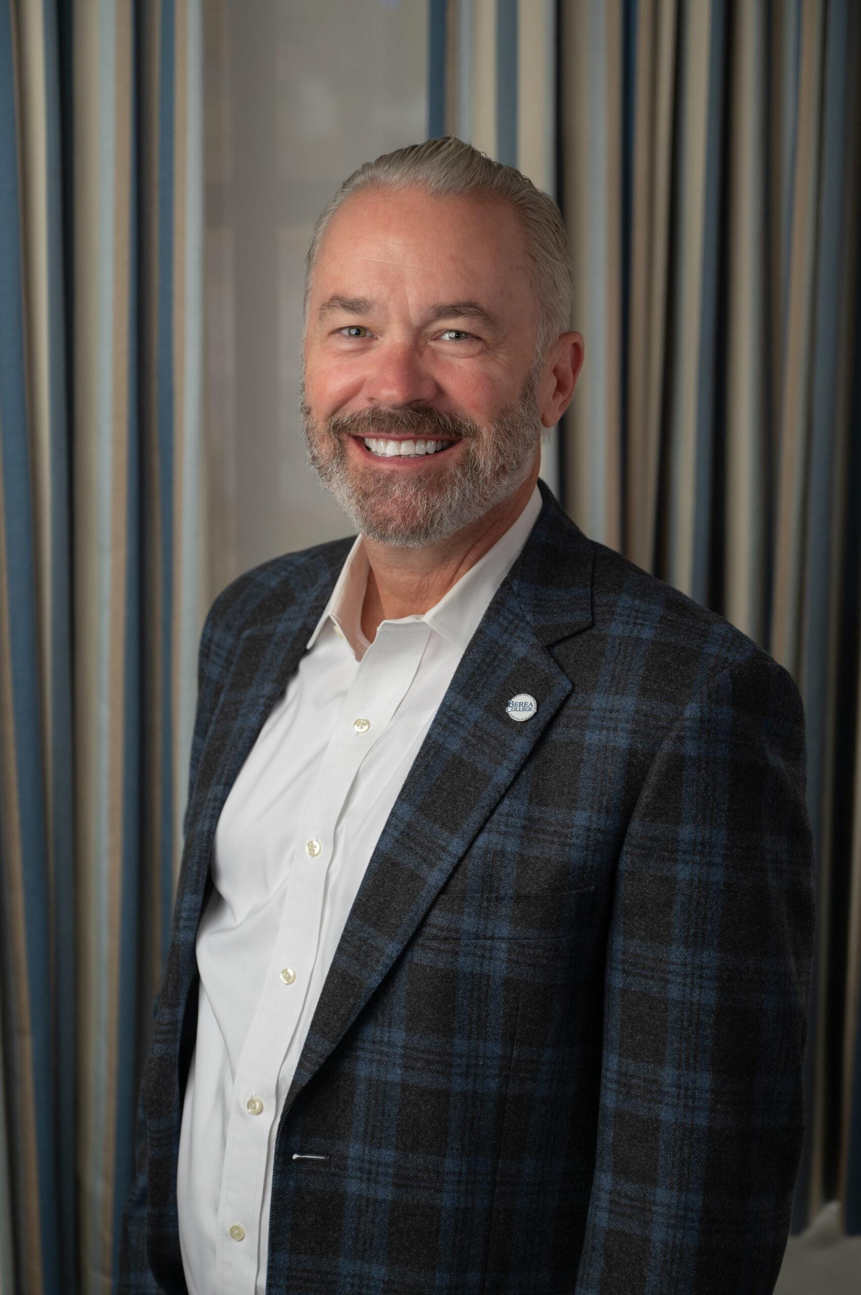 Chad Berry is standing in front of gray backdrop. He is smiling and wearing a blue and green checkered jacket with a white shirt.